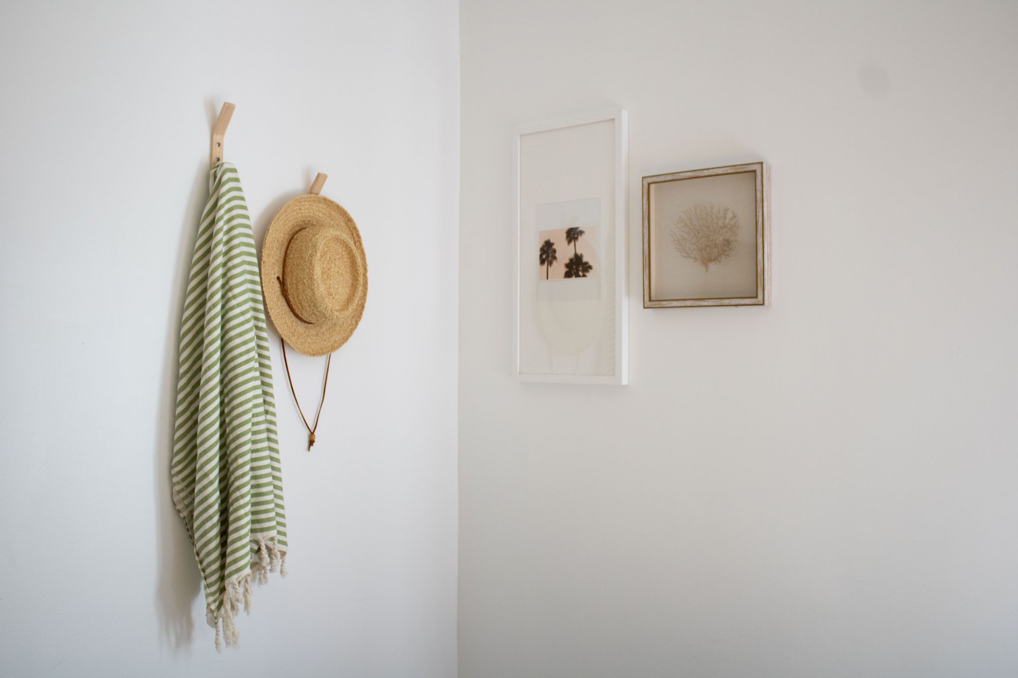 A minimalist entry wall with a green striped scarf, a straw hat on hooks, and two simple framed artworks on a white wall.