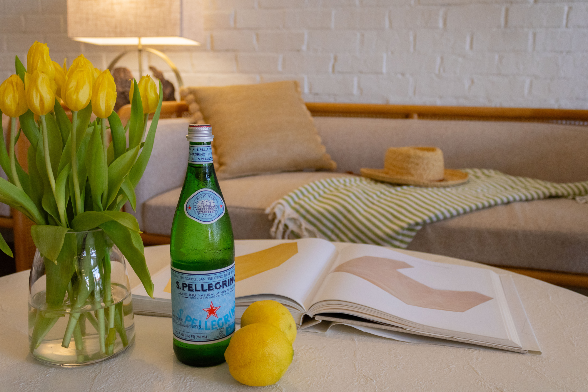 A cozy coffee-table scene: yellow tulips, a bottle of San Pellegrino, a lemon, an open magazine, and a straw hat on a striped throw.