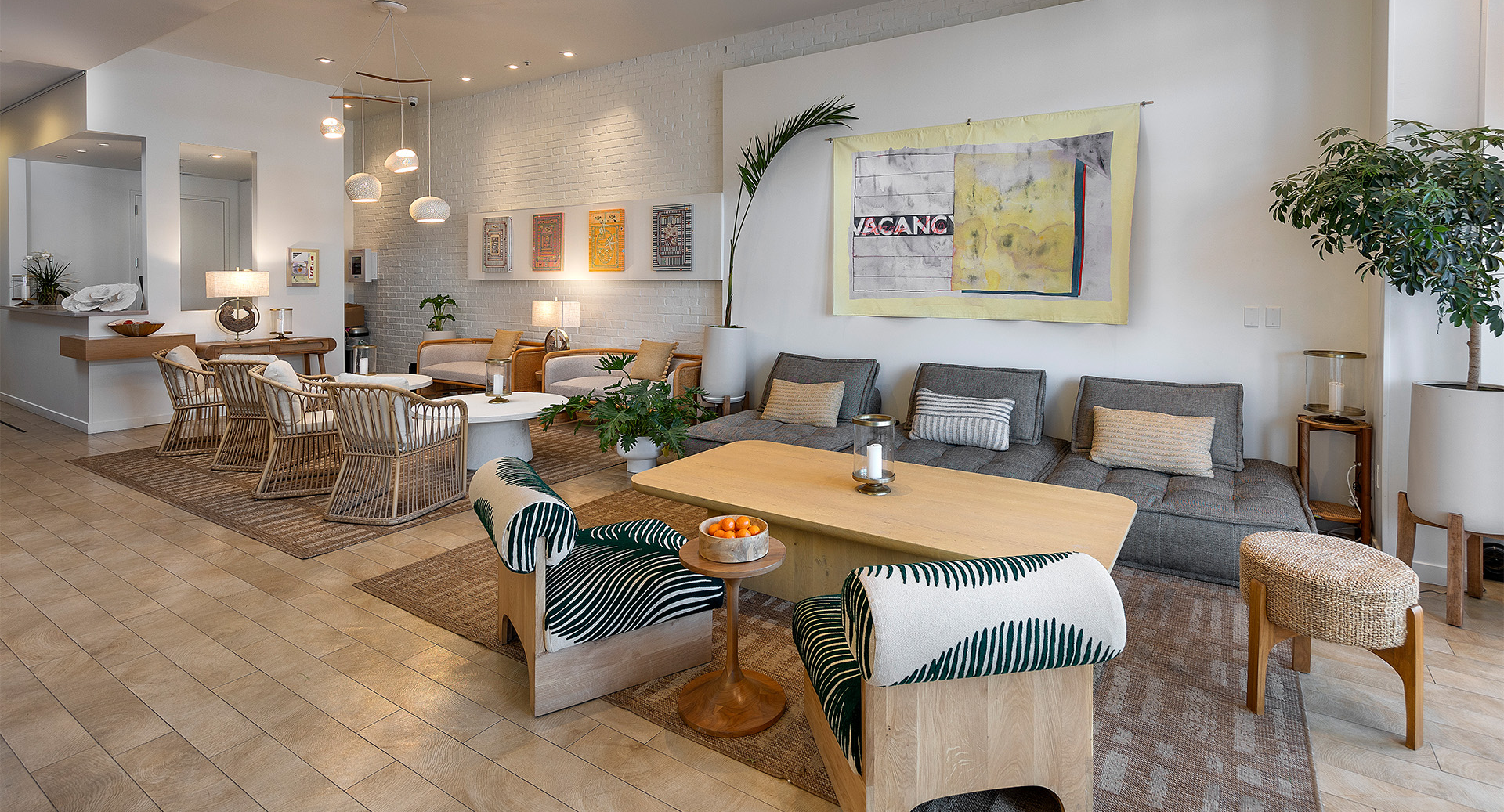A bright, modern living room with a large gray sofa, wooden coffee tables, woven chairs, plants, and art on the walls.