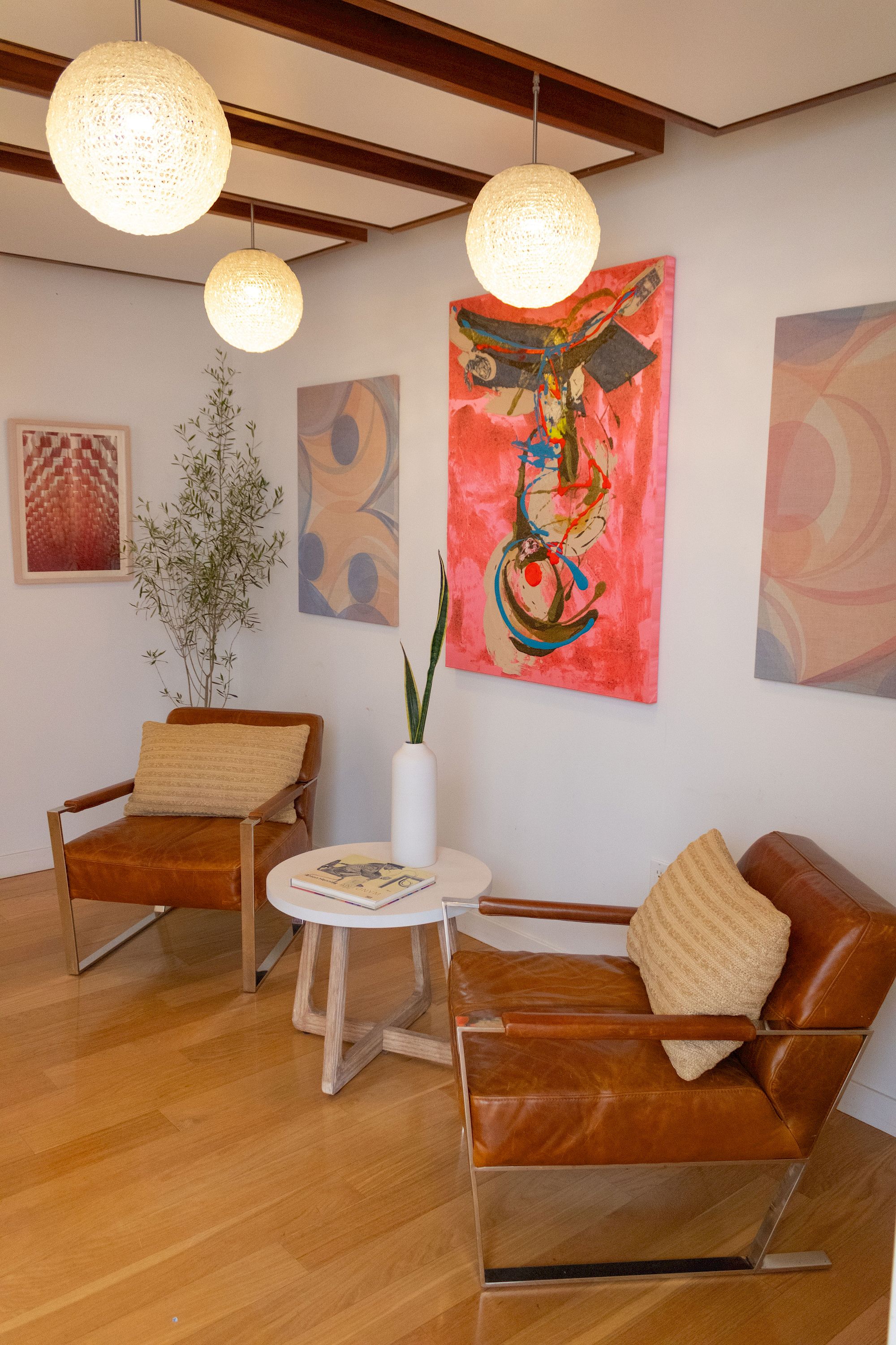 A cozy waiting area with mid-century chairs, a small round table, wall art, potted plant, and warm pendant lights, all in a bright, inviting space.