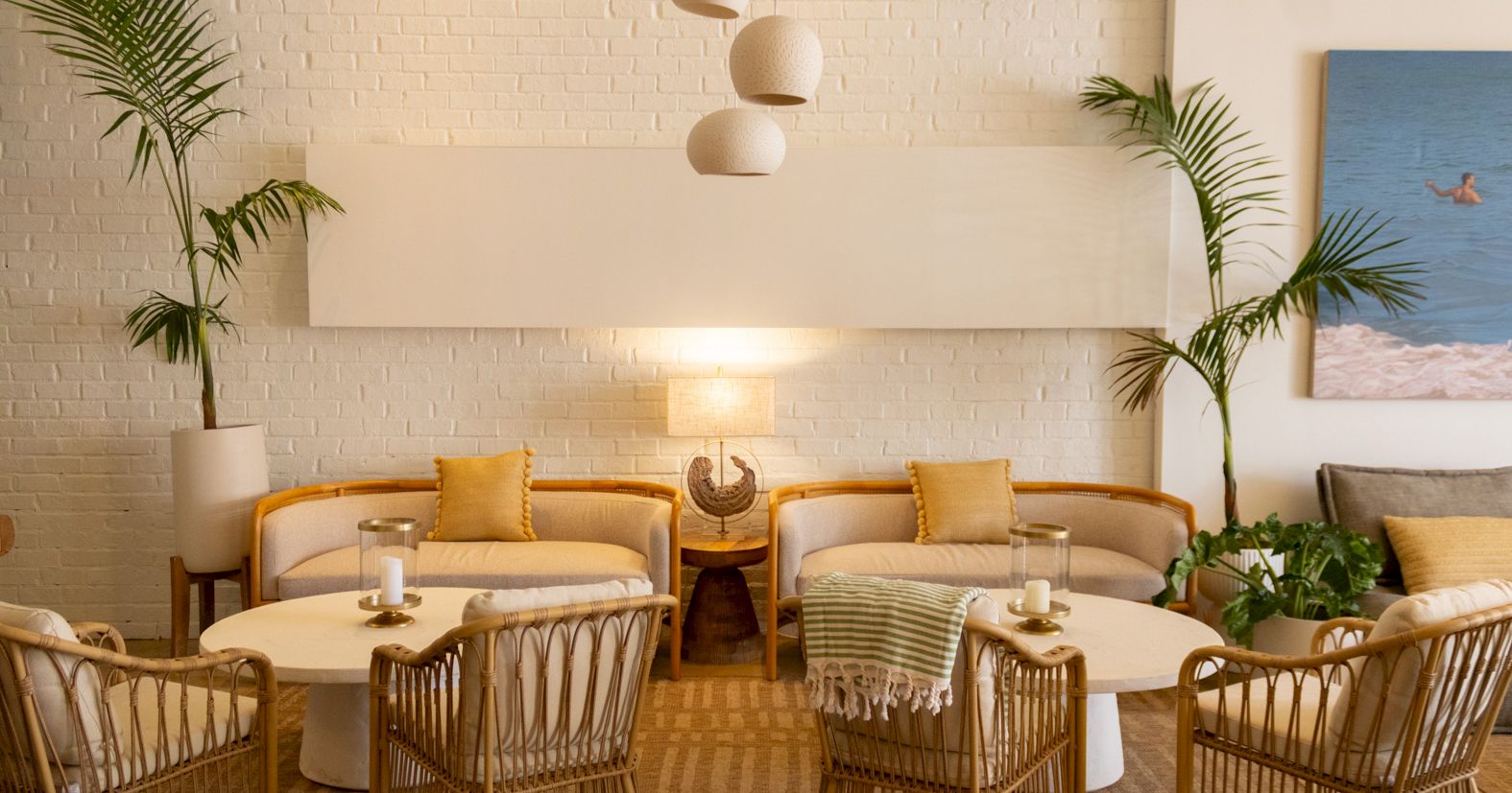 A stylish lounge with wicker chairs, round white tables, soft yellow cushions, potted palms, a hanging light feature, and warm ambient lighting.