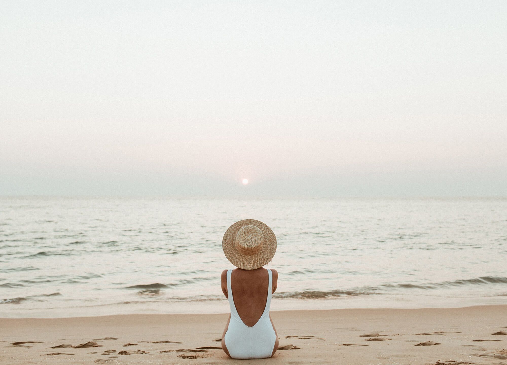 A person sits on the beach facing the ocean at sunset, wearing a white swimsuit and a wide-brimmed hat, as waves roll in.
