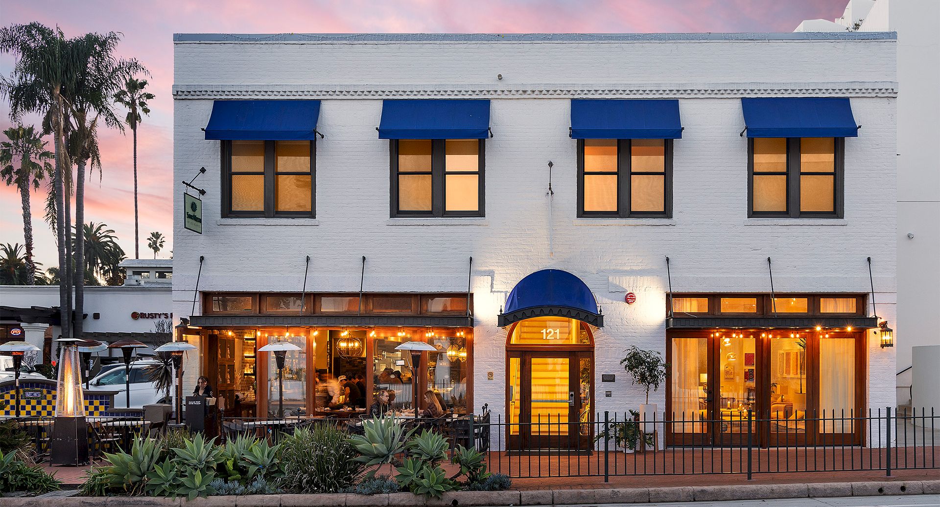 A white two-story building with blue awnings, warm-lit storefronts, outdoor seating, palm trees, and a pink sunset sky behind it.