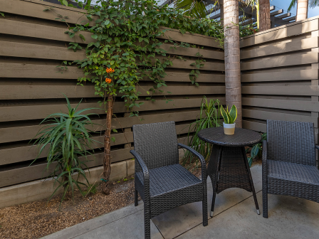 A small patio with two wicker chairs and a round table, potted plant, boardwood fence, and a tree in a cozy outdoor corner.