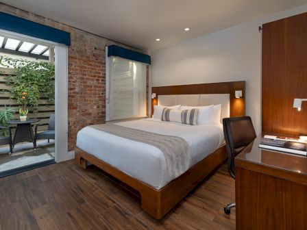 A modern hotel room with a wooden bed, white linens, a desk, and a balcony door opening to a small outdoor seating area.