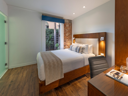 A stylish hotel room with a wooden bed, modern desk, soft lighting, brick accent wall, and a view of a small courtyard outside, creating a cozy, contemporary vibe.