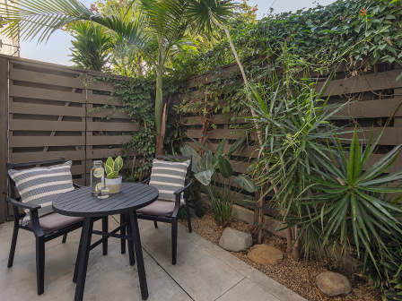 A small outdoor patio with a round table, four chairs with cushions, potted plants, and a wooden privacy fence surrounded by tropical greenery.