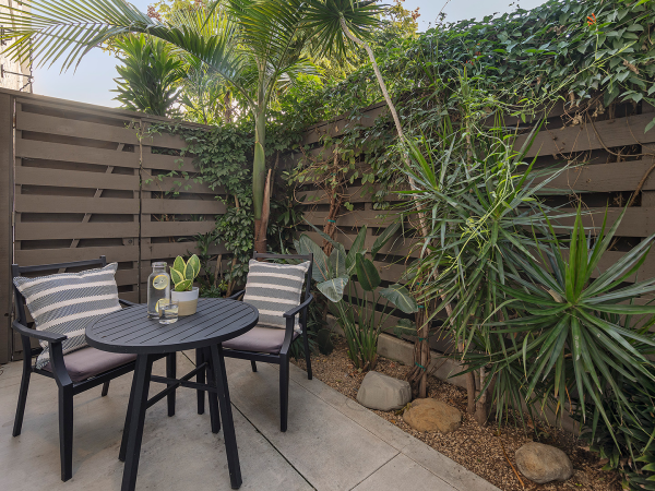 A small outdoor patio with a round table, four chairs with cushions, potted plants, and a wooden privacy fence surrounded by tropical greenery.
