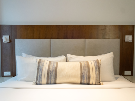 A neatly made bed with a padded gray headboard, wooden framing, and white pillows plus a striped lumbar pillow in a tidy hotel-style setup.