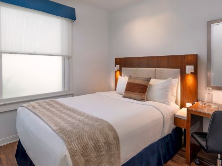 A tidy hotel room with a double bed, nightstands, lamp, desk, chair, wall-mounted TV, window with blinds, and a full-length mirror, neat and modern.