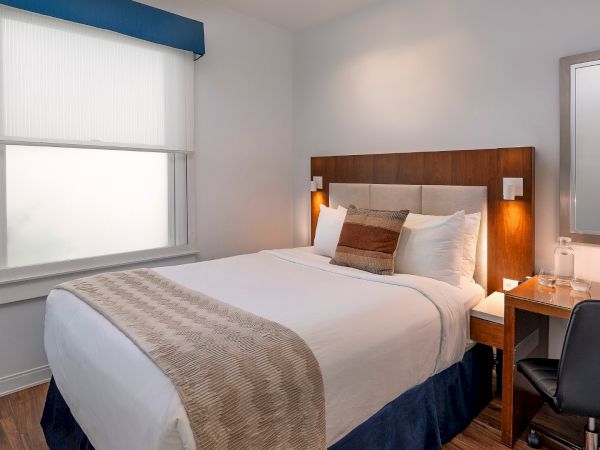 A tidy hotel room with a double bed, nightstands, lamp, desk, chair, wall-mounted TV, window with blinds, and a full-length mirror, neat and modern.