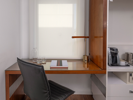 A small, modern home office with a wooden L-shaped desk, black swivel chair, lamp, window blind, and a nearby kitchenette with a coffee maker.