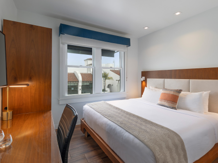 A modern hotel bedroom with a large bed, wooden headboard, two bedside tables, wall lamps, a desk with a chair, a wall-mounted TV, and a window with a blue valance.