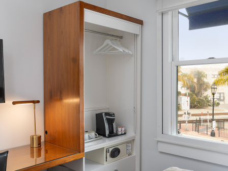 A hotel-style room with a desk, wall lamp, flat-screen TV, an open wardrobe with hangers, a coffee maker, safe, and a window looking out to palm trees. (140 chars, ends with a period.)