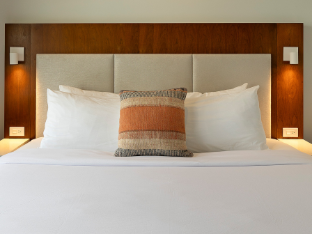 A neatly made hotel bed with white sheets, a central decorative pillow, and twin bedside tables with mounted lamps, in a wood-accented headboard.