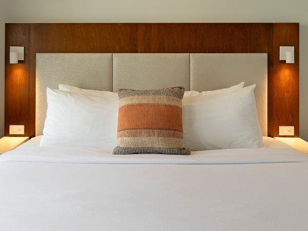 A neatly made hotel bed with white sheets, a central decorative pillow, and twin bedside tables with mounted lamps, in a wood-accented headboard.