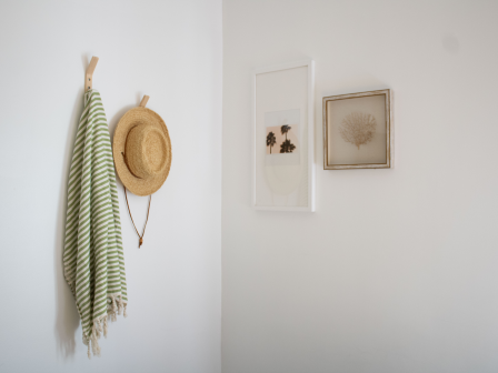 A minimal entryway scene with a striped green and white scarf, a straw hat hanging on a hook, and two framed wall art pieces on a white wall.