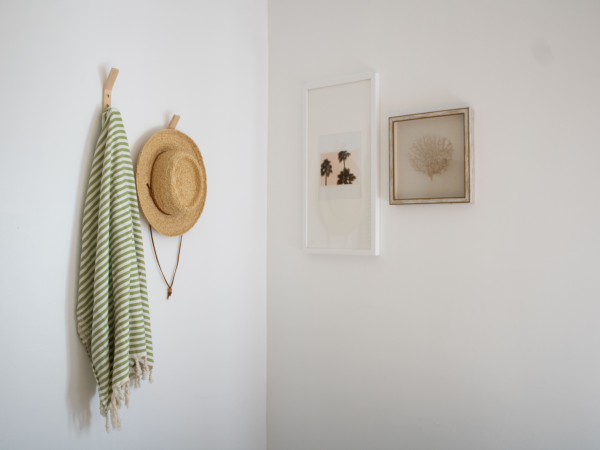 A minimal entryway scene with a striped green and white scarf, a straw hat hanging on a hook, and two framed wall art pieces on a white wall.