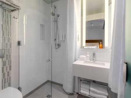 A modern white bathroom with a glass shower, tiled walls, a sink with storage below, a large mirror, and a white curtain on the right side.