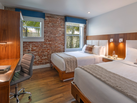 Modern hotel room with two queen beds, brick accent wall, wooden desk and chair, flat-screen TV, and warm bedside lighting. Clean and contemporary.