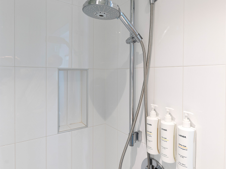 A modern shower with a rainhead, handheld wand, tiled walls, and three shampoo/conditioner bottles in a built-in niche.