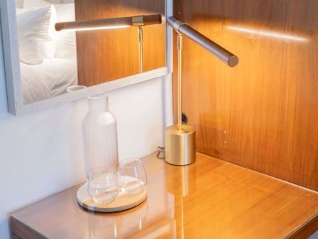 A wooden desk with a glass carafe, a metal lamp, and a mirror on the wall, reflecting a bed in the background, under warm light.