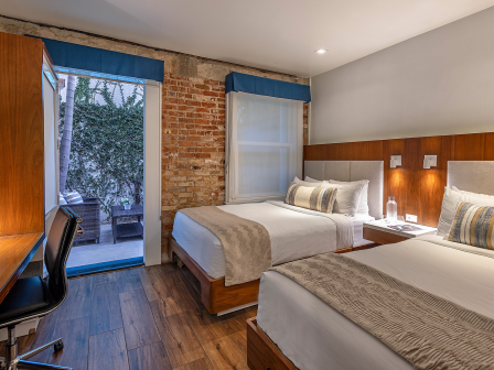 Two modern hotel beds with beige throws and striped cushions in a wood-paneled room, desk, chair, and a small balcony with brick wall view.