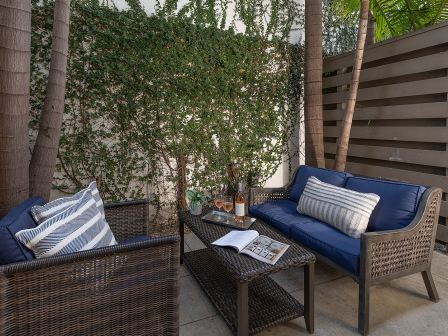 A cozy outdoor patio with wicker chairs, blue cushions, striped pillows, a glass coffee table, and a privacy fence among tall trees.