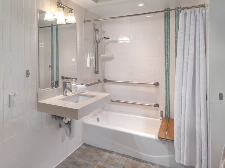 A compact, accessible bathroom with a white bathtub, wall-mounted sink, grab bars, a shower curtain, and a wooden bath seat by the tub.