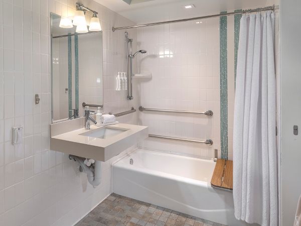 A compact, accessible bathroom with a white bathtub, wall-mounted sink, grab bars, a shower curtain, and a wooden bath seat by the tub.