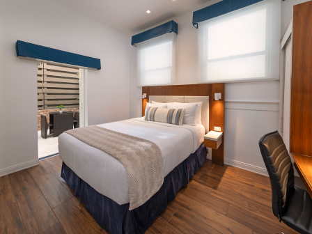 A modern hotel room with a queen bed, nightstands, desk and chair, wooden floor, blue window valances, and a doorway to a small seating area. (140 chars, ending with a period.)