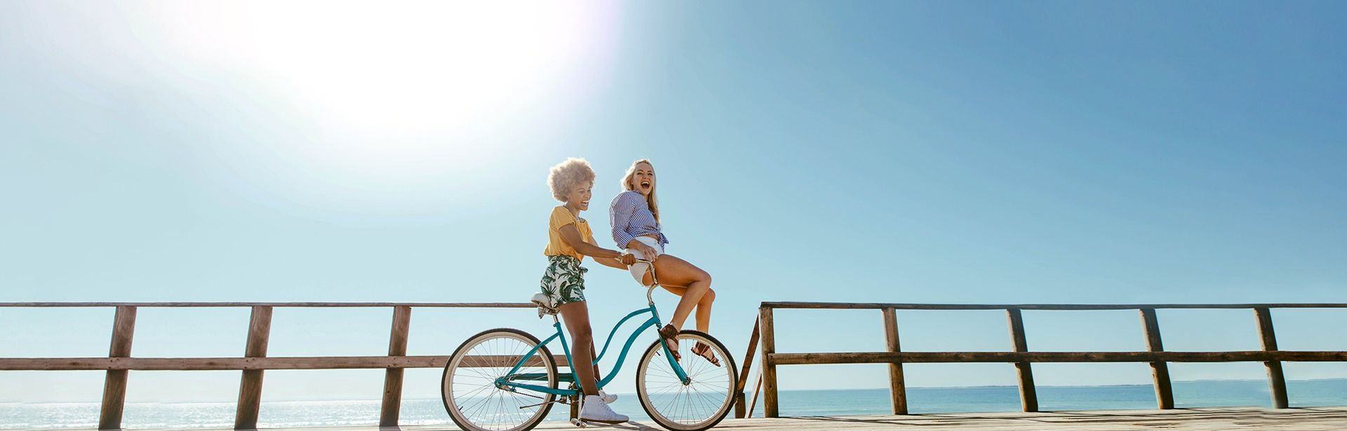 Two kids ride a teal bicycle on a sunny boardwalk, smiling as the ocean shimmers in the background.