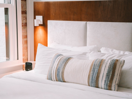 A neatly made hotel bed with white linens and a striped bolster pillow, beside a window and wall-mounted night light, in a modern room.
