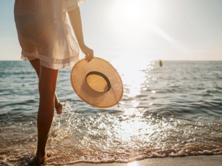 A person stands at the shore in a light dress, holding a wide-brimmed hat as the sun sets over calm waves. Ending with a period.