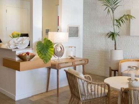 A bright, modern lobby or reception area with a wooden counter, decorative plants, wicker chairs, a beige sofa, and soft, neutral decor.