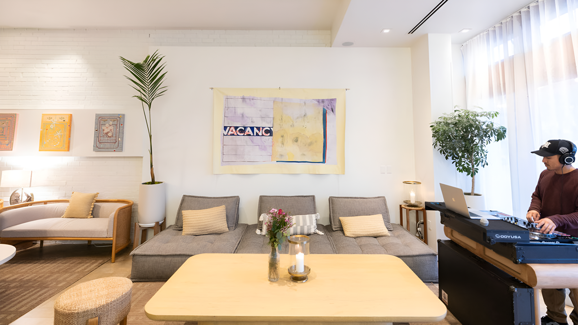 A bright, cozy lounge with a gray sofa, wooden coffee table, plants, soft art on the wall, and a DJ at a laptop setup in the corner.