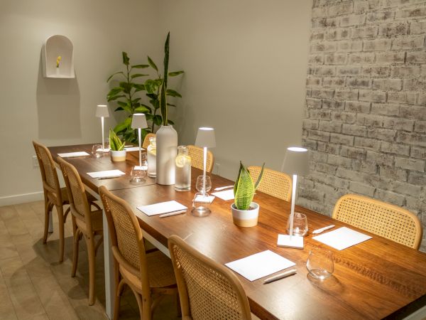 A long wooden dining table set with plates, cutlery, glasses, small plants, and pendant lamps in a modern, brick-walled restaurant.