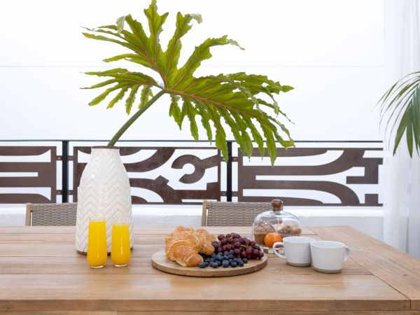 A bright outdoor breakfast setup with a wooden table, a big green plant, a vase with yellow juice, croissants, grapes, a glass cloche, and cups on a patterned railing backdrop.