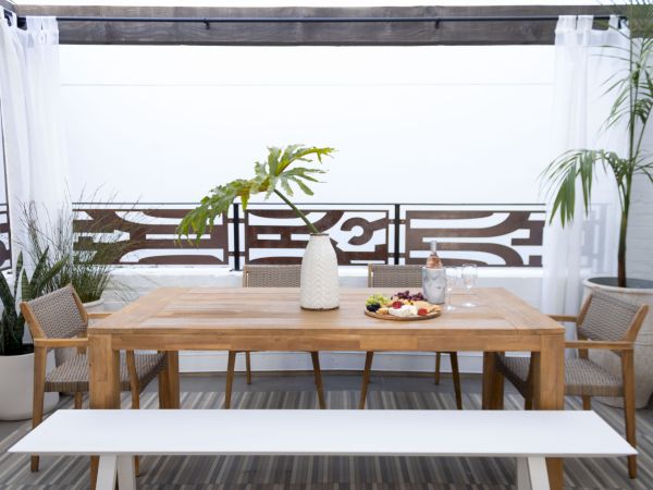 A cozy outdoor dining setup with a wooden table, two chairs, a white bench, potted plants, and a decorative railing on a sunny balcony.