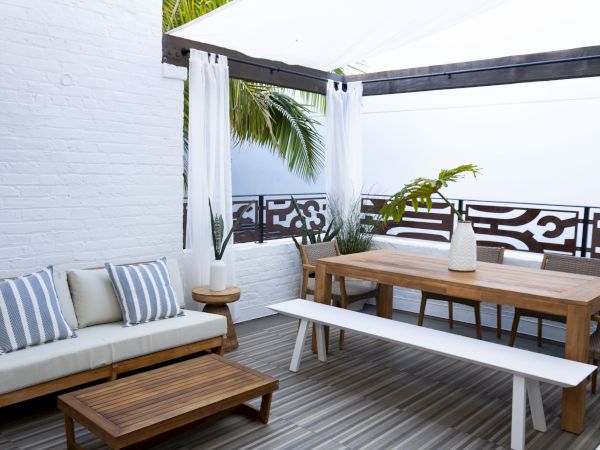A modern rooftop patio with a white-walled seating area, gray-striped cushions, a wooden dining table, white bench, and tropical potted plants by a decorative railing.