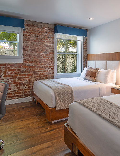 A modern hotel room with two queen beds, a brick feature wall, wooden desk and chair, a TV, and bedside lamps, plus a cozy throw and cushions ending.