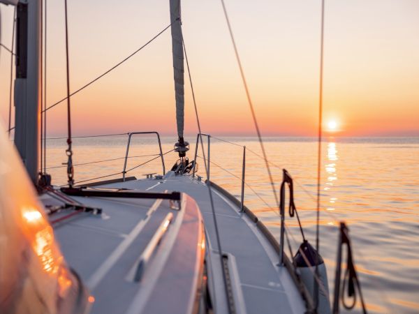 Sunset over calm sea from a sailboat deck, lines and rails fade into warm orange light; tranquil voyage moment on tranquil waters.