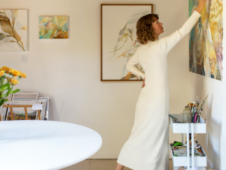 A woman in a white dress hangs or arranges abstract art on a gallery wall, while a vase of flowers sits on a round table in the foreground.