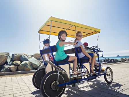 A group of four people riding a yellow-topped pedal-powered quad bike on a sunny seaside promenade.