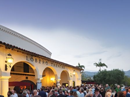 A crowd gathers outside the Lobero Theatre at dusk, people chatting under warm lights near arched entrances and a pale, clear sky.