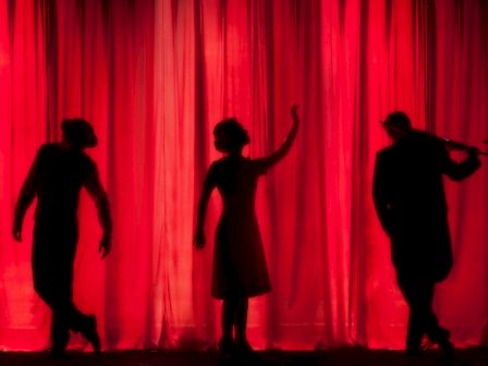 Three silhouetted performers against a red curtain, posing with dynamic gestures on stage.