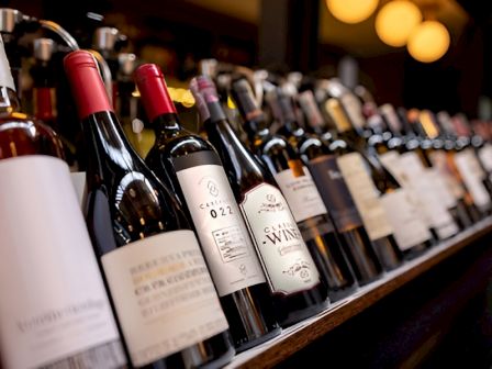 A row of assorted wine bottles on a bar shelf, angled toward the camera, labels facing outward, warm lighting in the background.