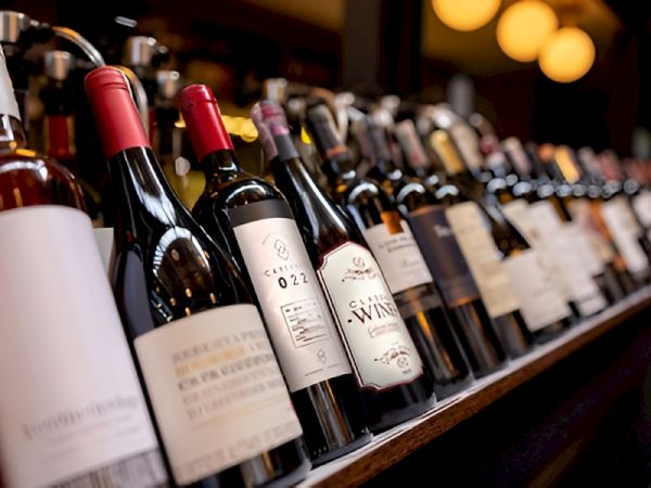 A row of assorted wine bottles on a bar shelf, angled toward the camera, labels facing outward, warm lighting in the background.