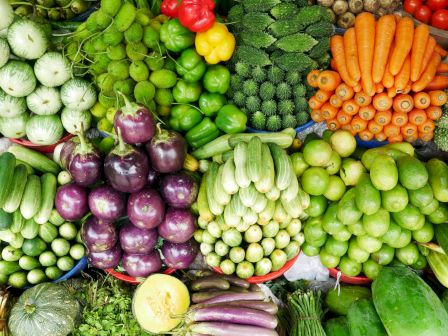 A colorful display of fresh vegetables: onions, peppers, carrots, greens, cucumbers, eggplants, broccoli, cabbage, and leafy greens in vibrant rows.