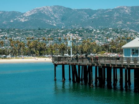 A wooden pier extends into calm blue water with a seaside town, palm trees, and mountains in the background, under a bright sky.